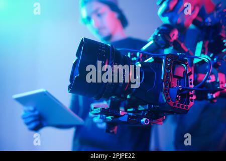Primo piano dell'operatore che utilizza una fotocamera professionale durante le riprese con il regista che controlla il processo Foto Stock