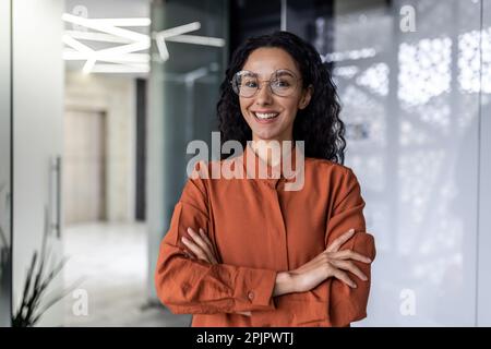 Giovane donna d'affari ispanica bella e di successo sorridente e guardando la macchina fotografica, lavoratrice che lavora all'interno dell'ufficio indossando occhiali e capelli ricci, braccia incrociate ritratto. Foto Stock