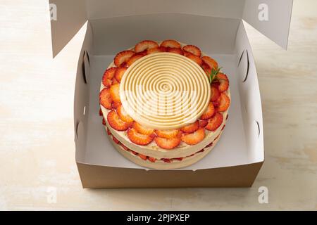 Torta di compleanno vegana con vaniglia e fragola ricoperta di cioccolato bianco in una scatola di cartone aperta su un tavolo e uno sfondo bianco Foto Stock