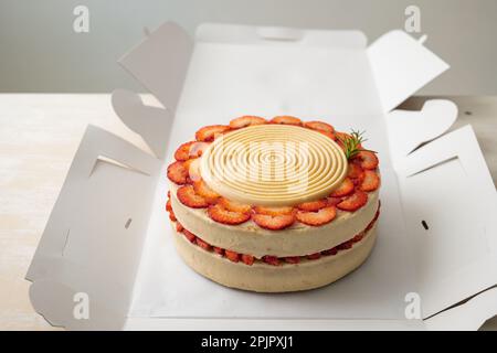 Torta di compleanno vegana con vaniglia e fragole ricoperte di cioccolato bianco in una scatola di cartone aperta su un tavolo e uno sfondo bianco Foto Stock