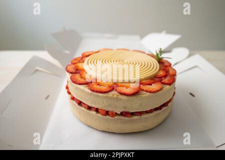 Torta di compleanno vegana con vaniglia e fragola ricoperta di cioccolato bianco in una scatola di cartone aperta su un tavolo e uno sfondo bianco Foto Stock