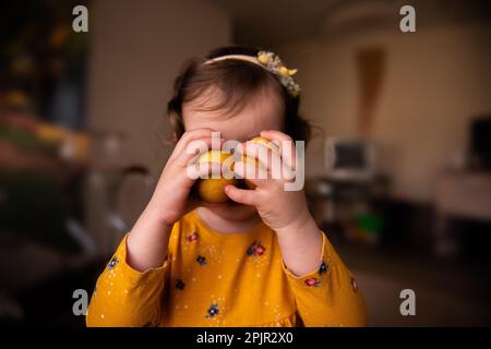 La bambina in vestito contiene due uova di Pasqua dipinte con vernice naturale curcuma vicino al viso. Decorazione casa in vacanza luminosa. Atmosfera di stile di vita. Che Foto Stock