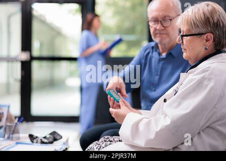 Medico senior che prende il test del livello di insulina del paziente usando il glucometro nell'area di attesa dell'ospedale, facendo l'esame del diabete per misurare il glucosio. Uomo anziano con visita di controllo Foto Stock