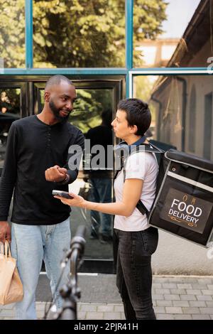 Cliente afro-americano che paga per la consegna di cibo per ufficio con Smart Watch. Donna corriere con dispositivo terminale POS, cliente che effettua il pagamento senza contatto per il pasto da asporto Foto Stock