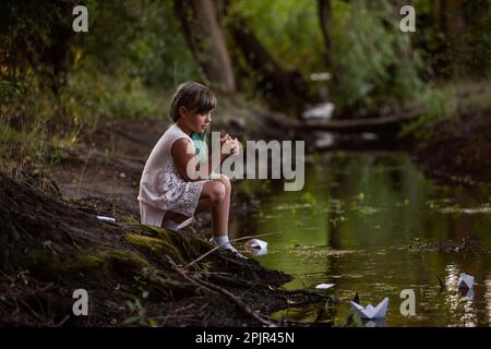 Ragazza adolescente con capelli verdi in vintage vestito, si trova sulla riva del fiume, lancia carta bianca origami barca in acqua nella foresta. Traslazione concep Foto Stock