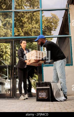Corriere che passa il cliente scatole pizza stack, uomo che prende l'ordine da zaino termico vicino all'edificio degli uffici all'aperto. Servizio di consegna cibo, pizzeria africano americano consegna deliveryman holding takeaway Foto Stock