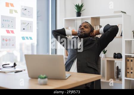 Uomo esausto in tuta con le mani attaccate dietro la testa mentre si è seduti alla scrivania con un computer portatile. Dipendente afro-americano che lotta contro la stanchezza causata da lunghe ore di lavoro in ufficio moderno. Foto Stock