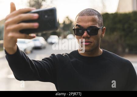Uomo con capelli corti e abiti casual con occhiali da sole che porta il selfie con uno smartphone moderno all'aperto. Sfondo sfocato. Foto di alta qualità Foto Stock