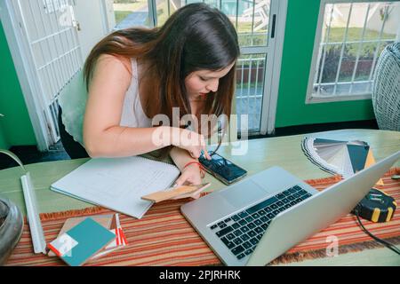 giovane donna ispanica latina interior designer invia una nota vocale per telefono al suo cliente, lavorando a casa, appoggiandosi sul tavolo lavorando con il portatile, Foto Stock