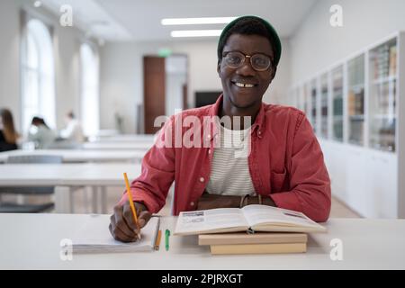 Giovane afroamericano che studia in biblioteca universitaria prendendo appunti in blocco note. Foto Stock