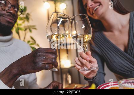 Un primo piano di bicchieri da vino come coppia interrazziale - l'uomo africano e la donna caucasica - brinda con vino bianco, i loro volti sfocati ridendo nel bac Foto Stock