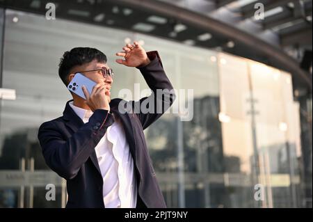 Un uomo d'affari asiatico o un capo maschile di successo e fiducioso in una tuta aziendale formale parla al telefono con il suo business partner mentre standi Foto Stock