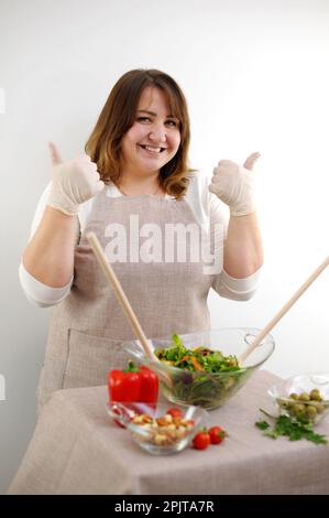 Allegro chef femminile è seduto al tavolo con un mazzo di verdure, mostrando il pasto preparato e ok segno.Female chef mostrando il pasto preparato e ok segno di alta qualità foto Foto Stock