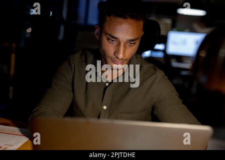 Uomo d'affari biraciale seduto alla scrivania, con un computer portatile e lavorando tardi in ufficio Foto Stock