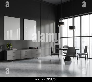 Interno di elegante sala da pranzo con pareti grigie, pavimento in cemento, tavolo rotondo con sedie e comò grigio con due poster verticali. Finestra wi Foto Stock