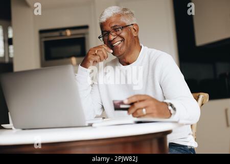 Allegro uomo anziano sorridente alla macchina fotografica mentre acquista in linea a casa. Uomo maturo felice che usa la sua carta di credito per effettuare un ordine online su un computer portatile. Foto Stock