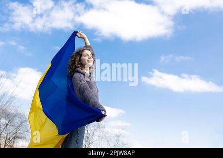 Una ragazza adulta di 18 anni, ha una bandiera Ucraina giallo-blu dietro la schiena contro il cielo, guarda avanti, nella speranza di un futuro più luminoso. Ukra Foto Stock