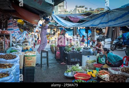 Una donna si alza su uno sgabello per regolare un baldacchino in un'area del mercato centrale in PLEIKU, Vietnam, che vende frutta, verdura e prodotti secchi. Foto Stock