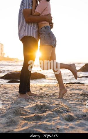 A metà di una coppia felice e varia che si abbraccia in spiaggia durante il tramonto Foto Stock