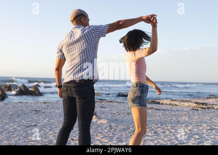 Felice coppia diversa tenendo le mani e ballando insieme in spiaggia Foto Stock