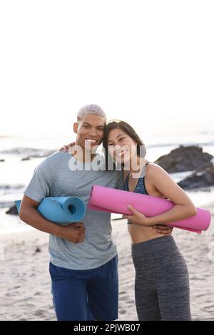 Ritratto di coppia felice e variegata indossando abbigliamento sportivo, tenendo stuoie da yoga e sorridendo in spiaggia Foto Stock