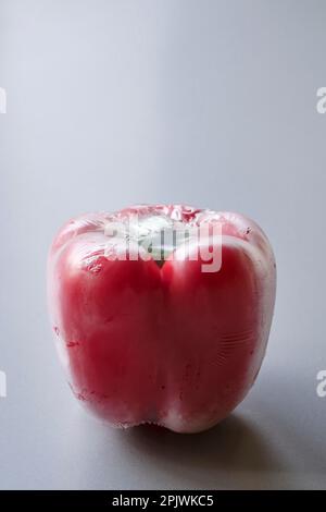 Peperone rosso sigillato in involucro di plastica su fondo grigio. Immagine verticale drammatica di una paprika di un supermercato confezionata in foglio di plastica. Foto Stock