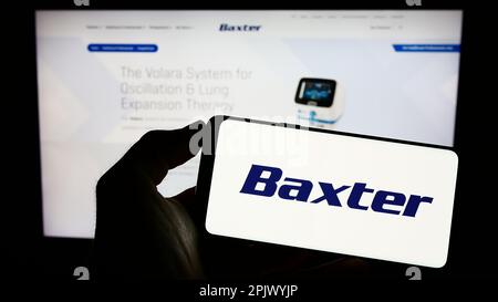 Persona che tiene il telefono cellulare con il logo della società sanitaria americana Baxter International Inc. Sullo schermo di fronte alla pagina web. Messa a fuoco sul display del telefono. Foto Stock