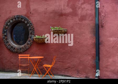 Un luogo invitante dove sedersi contro questo muro di mattoni rosso intonacato e una finestra rotonda, con Pansie in vaso e sedie in metallo arancione e tavolo. Foto Stock