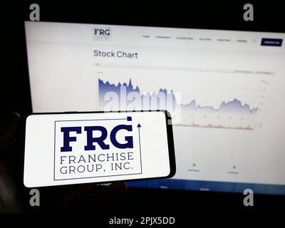 Persona in possesso di un telefono cellulare con il logo della società americana Franchise Group Inc. (FRG) sullo schermo davanti alla pagina Web. Messa a fuoco sul display del telefono. Foto Stock