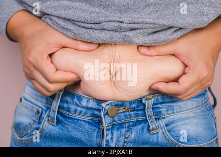 Mano di donna grassa che tiene grasso eccessivo della pancia isolato su sfondo rosa. Pancia grassa in sovrappeso della donna. Dieta femminile e concetto di salute del corpo Foto Stock