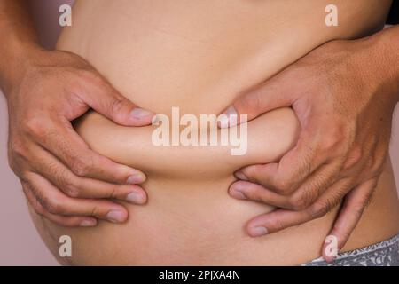 Mano maschile grassa che tiene eccessivo grasso della pancia isolato su sfondo rosa. L'uomo tocca il ventre grasso. Giovane uomo in sovrappeso con grande pancia Foto Stock