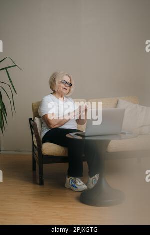 Donna anziana che applica crema cosmetica sulle mani mentre lavora lontano da casa utilizzando un dispositivo portatile, sorridendo allegro. Concetto di bellezza. Persone Foto Stock