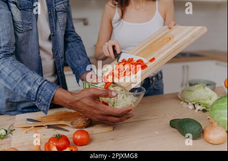 Primo piano di un tagliere con le mani di una coppia che taglia insieme le verdure in cucina. Cibo vegetariano sano Foto Stock
