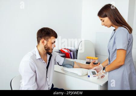 Infermiere in uniforme medica e guanti trasparenti per il controllo della pressione sanguigna di un giovane uomo in abiti casual. La donna della brunetta preme il tasto sul tonometro. Te Foto Stock