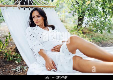 Bella donna incinta si trova in amaca nel giardino. Giovane brunetta femminile in attesa di bambino in abito bianco riposante all'aperto. Alberi verdi in estate Foto Stock