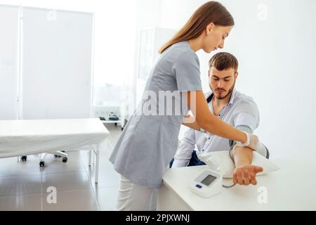 Infermiere in uniforme medica e guanti trasparenti per il controllo della pressione sanguigna di un giovane uomo in abiti casual. La donna della brunetta preme il tasto sul tonometro. Te Foto Stock