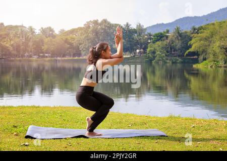 Donna asiatica che pratica yoga facendo Eagle posa sul tappeto accanto a un lago nel parco all'aperto. Foto Stock