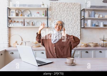 Giovane studentessa latino-americana in hijab e cuffie che studiano a casa di fronte al portatile, mani in su, ballare, ascoltare la lezione. Sorridendo, guarda la fotocamera. Foto Stock