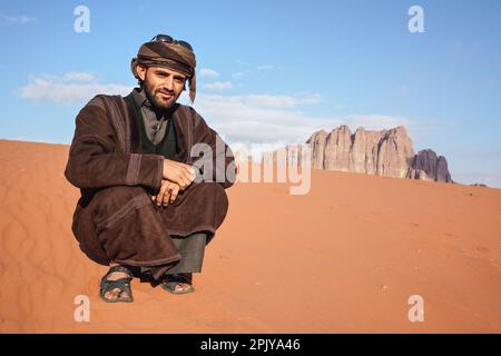 Wadi Rum, Giordania - 19 gennaio 2020: Sconosciuto uomo arabo in tradizionale bisht beduino cappotto caldo accovacciato nel deserto di sabbia rossa nel giorno di sole - montagne bac Foto Stock