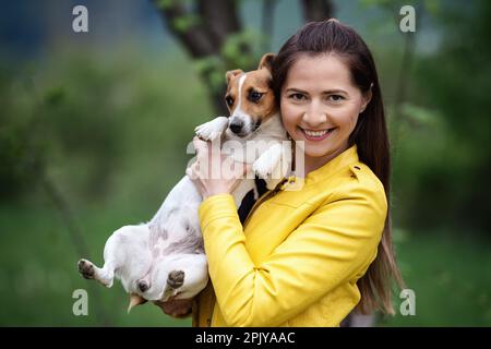 Giovane donna in giacca gialla che tiene il suo cane Jack Russell terrier sulle mani, sfondo di alberi verdi sfocati Foto Stock