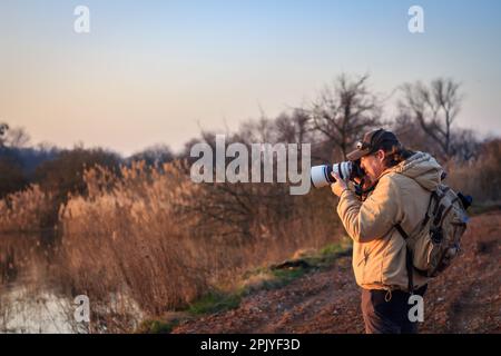 Fotografo di paesaggi con fotocamera e teleobiettivo che fotografa la fauna selvatica durante il tramonto Foto Stock