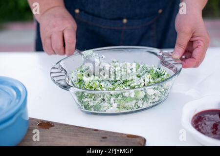 Vecchia cuoca femminile che lavora all'aperto in estate, tenendo il cucchiaio, mescolando insalata con formaggio. Foto Stock