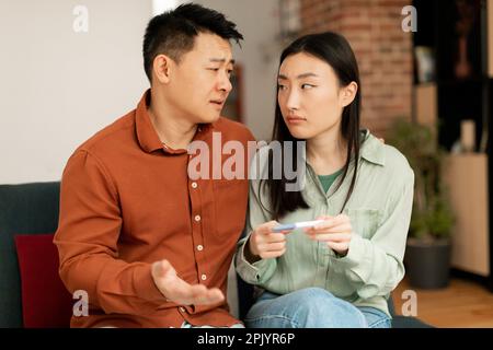 Depresso triste marito asiatico e moglie sconvolto di risultato negativo test di gravidanza, seduta sul divano in soggiorno interno Foto Stock
