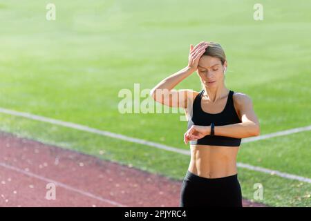 Atleta maturo stanco e sconvolto che controlla la frequenza cardiaca sul bracciale fitness Smart Watch, runner in stadio dopo esercizio attivo e jogging nelle giornate di sole, donna bionda in abbigliamento sportivo. Foto Stock