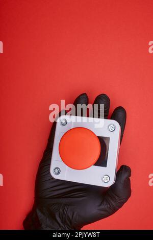 La mano nel guanto in vinile nero, che tiene il pulsante di emergenza sullo sfondo rosso nella vista dall'alto Foto Stock