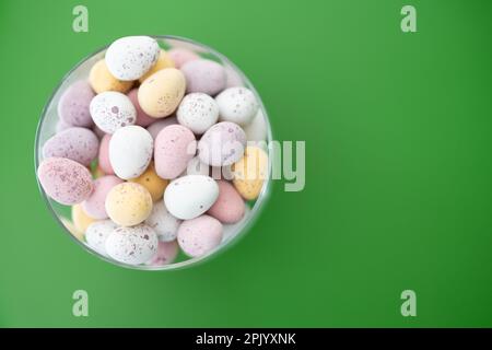 Mini uova al cioccolato rosa, giallo, bianco e viola in un recipiente di vetro su sfondo verde (Pasqua) Foto Stock