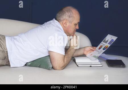 Un uomo d'affari si trova su un divano e guarda le tendenze dei dati su grafici e grafici. Lavoro a distanza Foto Stock