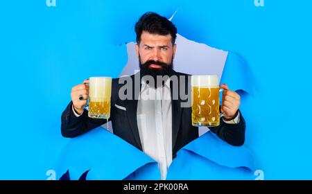 Tempo di birra. Uomo barbuto con tazza di birra artigianale guardando attraverso il foro di carta. Pubblicità bar o pub. Alcool. Birra artigianale in Germania. Uomo d'affari in tuta Foto Stock