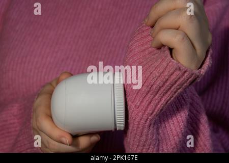 Donna che usa un rasoio Anti-pilling a casa. Dispositivo per la rasatura degli abiti. Rasoio in tessuto anti-peluche. Pulitore elettrico portatile pillola defuzzer pelucchi da rosa acrilico o lana maglione. Moda lenta che ripara i vestiti Foto Stock