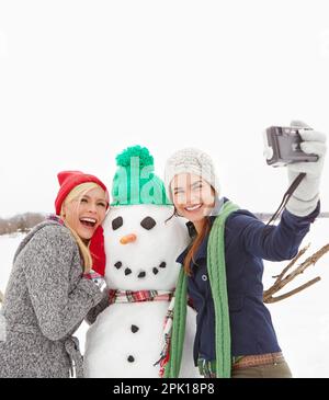 Buone vacanze da tre migliori amici. due bellissime giovani donne che prendono un selfie con un pupazzo di neve. Foto Stock